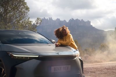 Person lehnt auf der Motorhaube eines SUVs und lächelt. Im Hintergrund ist eine bergige Landschaft zu erkennen.