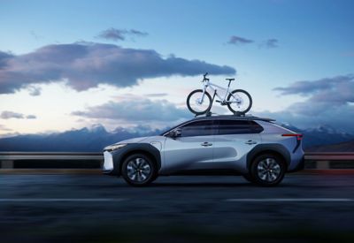Seitenansicht eines fahrenden, silbergrauen Toyota bZ4X mit auf dem Dach montierten Mountainbike, im Hintergrund schneebedeckte Berge