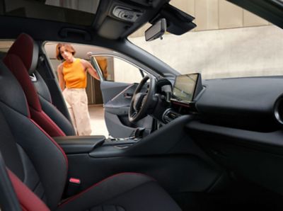Innenraum eines SUV-Modells  von der Beifahrerseite mit schwarzen Sportsitzen und roter Ziernaht, während eine Frau die Fahrertür öffnet.