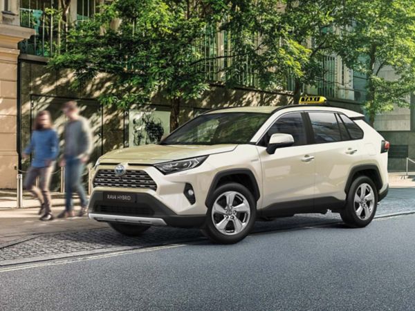 Ein beigen RAV4 Hybrid mit einem Taxi Schild auf dem Dach steht an einer Stadtstraße vor grünen Bäumen und einer Gebäudefassade.