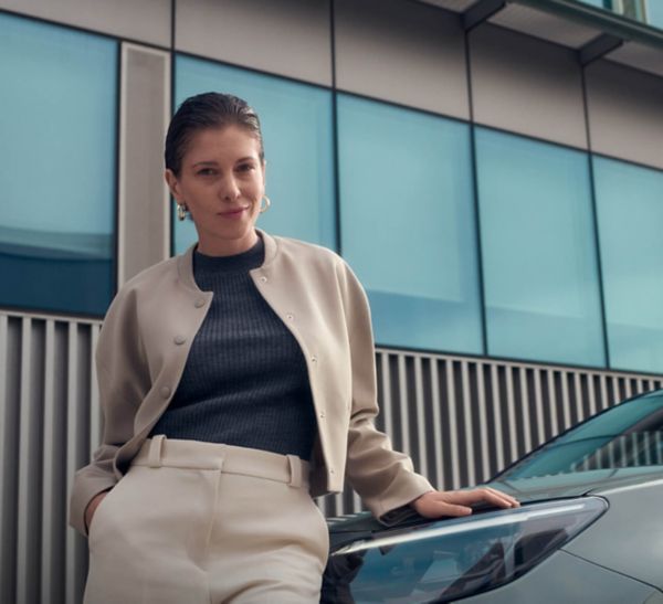Junge Frau mit schlichter Frisur und eleganter Kleidung lehnt entspannt an einem Auto, vor einer modernen Glasfassade.
