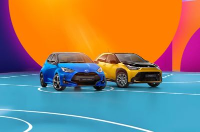 Blauer Toyota Yaris und gelber Toyota Aygo X auf einem türkisfarbenen Basketballfeld vor farbenfroher Architektur mit großem orangefarbenem Kreis.