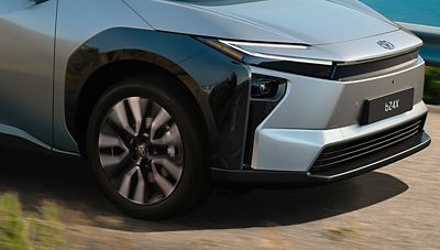 Nahaufnahme der Front und des Vorderrads eines silbergrauen Toyota bZ4X in Fahrt auf einer Straße nahe einem Gewässer.