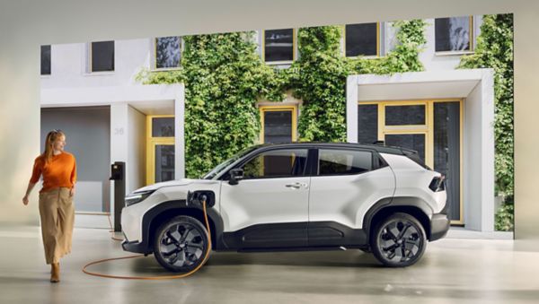 Vor einer Hauskulisse steht ein weißer Elektro SUV angeschlossen an einer Ladesäule und ein Person entfernt sich gerade davon.