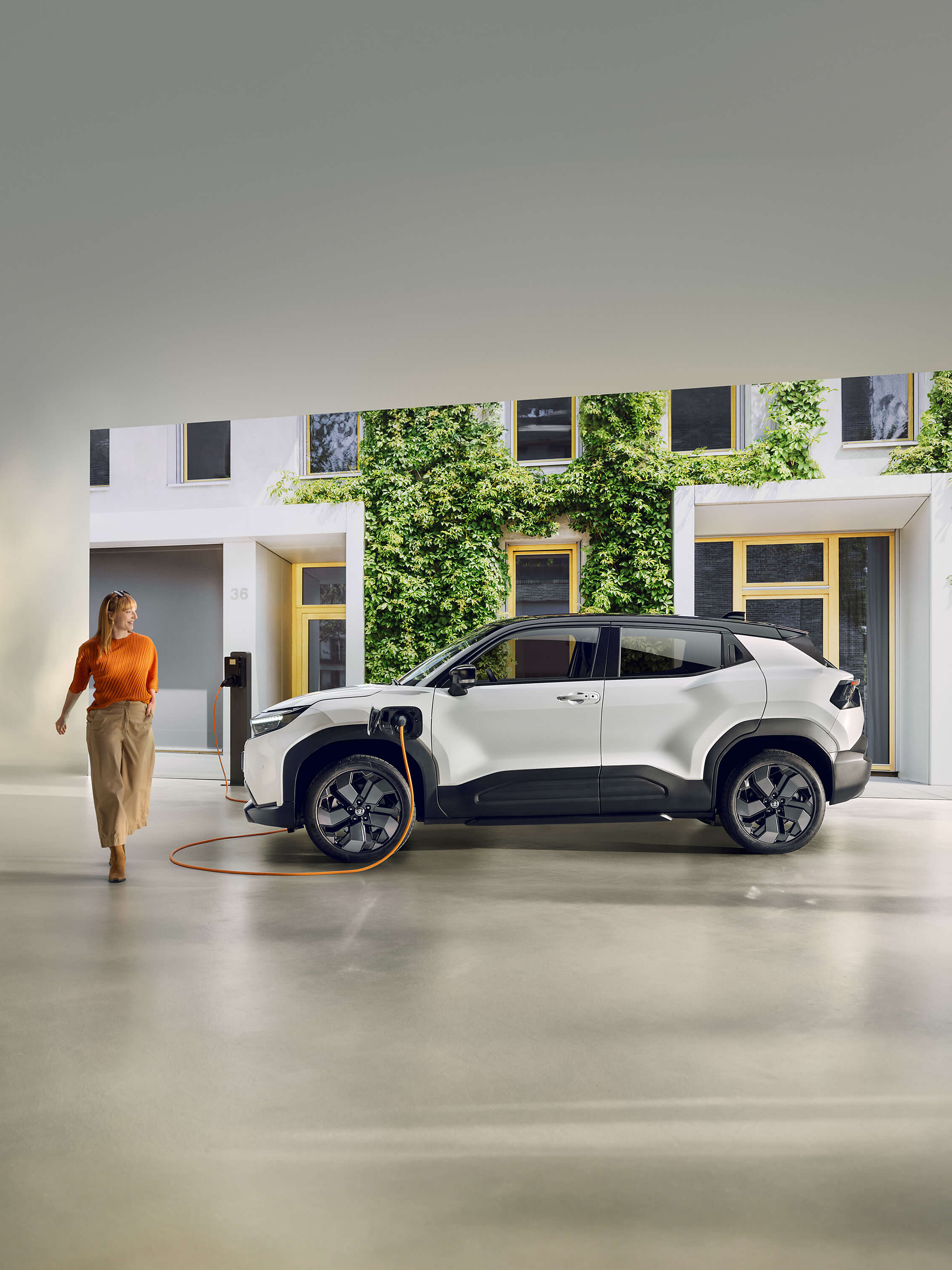 Ein schwarz-weißer Toyota Urban Cruiser steht vor einer weißen Hausfassade, die mit grünem Moos bewachsen ist, während ein orangenes Kabel das Auto über eine Wallbox lädt. Vom Auto weg läuft eine junge, lächelnde Frau.