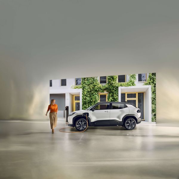 Ein schwarz-weißer Toyota Urban Cruiser steht vor einer weißen Hausfassade, die mit grünem Moos bewachsen ist, während ein orangenes Kabel das Auto über eine Wallbox lädt. Vom Auto weg läuft eine junge, lächelnde Frau.