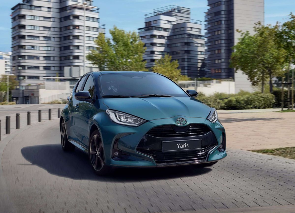 Blauer Toyota Yaris fährt auf einer Straße in der Stadt, dahinter sind Wohngebäude zu sehen.