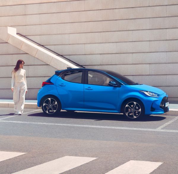 Eine Person läuft am Heck eines blauen Toyota Yaris entlang, der an der Seite geparkt. Die Farbgebung ist sehr eintönig, nur die blaue Lackierung sticht hervor.