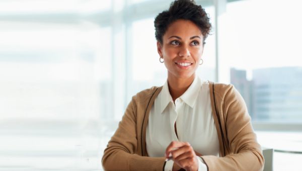 eine Frau sitzt in einem Büro, im Hintergrund sind Hochhäuser zu sehen, und lächelt