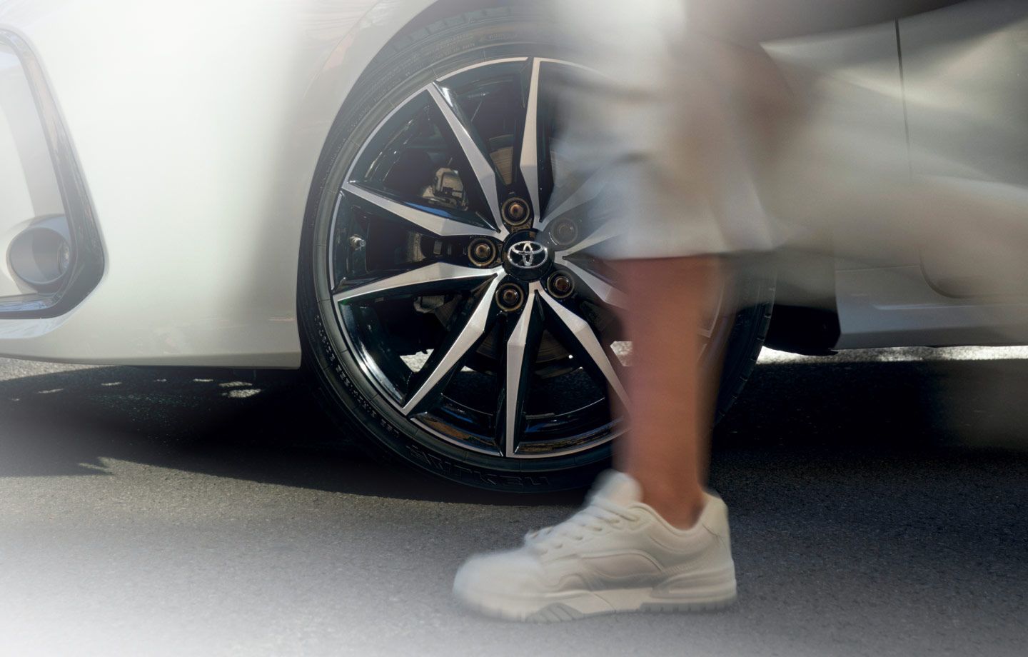 Nahaufnahme von Felgen eines weißen Autos, während im Vordergrund eine Person mit weißen Sneakern an dem Auto vorbeiläuft.