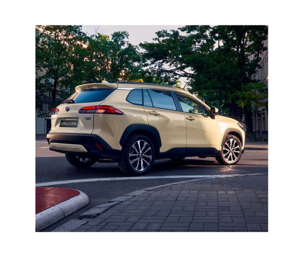 Ein beiger Corolla Cross Hybrid, der auf einer Straße in der Stadt geparkt ist mit grünen Bäumen im Hintergrund.