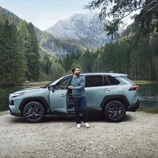 Mężczyzna wysiada z Toyoty nad jeziorem w lesie i podziwia widok, w tle góry