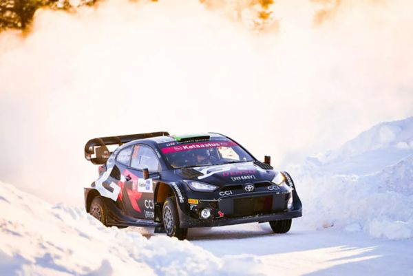 Ein schwarzes Rallye-Auto, das eine verschneite Strecke entlangfährt und dabei Schnee und Staub aufwirbelt, während das Sonnenlicht die Szene erhellt. Das Auto ist mit einem auffälligen Logo und Rennaufklebern versehen.