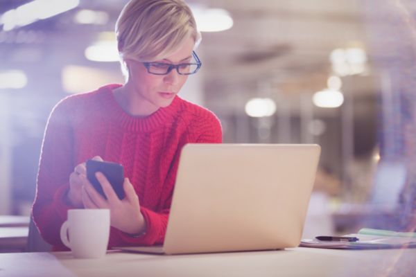 En kvinna med en röd tröja och glasögon sitter vid sin laptop med sin mobiltelefon i handen. Bredvid datorn står en kaffekopp.