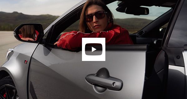 Nő napszemüvegben, piros dzsekiben, egy ezüst Toyota sportautó ablakán kihajolva, hegyes táj előtt.